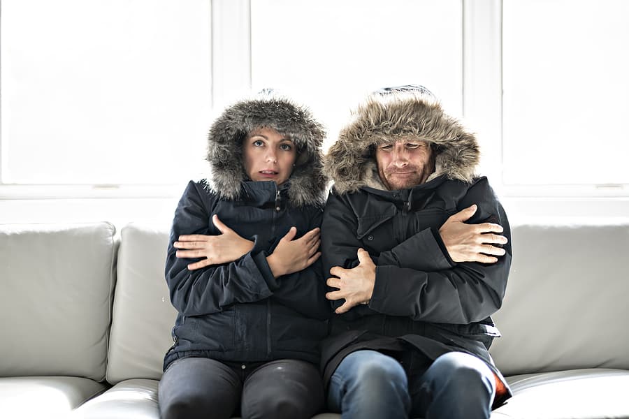 Heating Repair Service. Couple sitting cold inside house with winter coats.